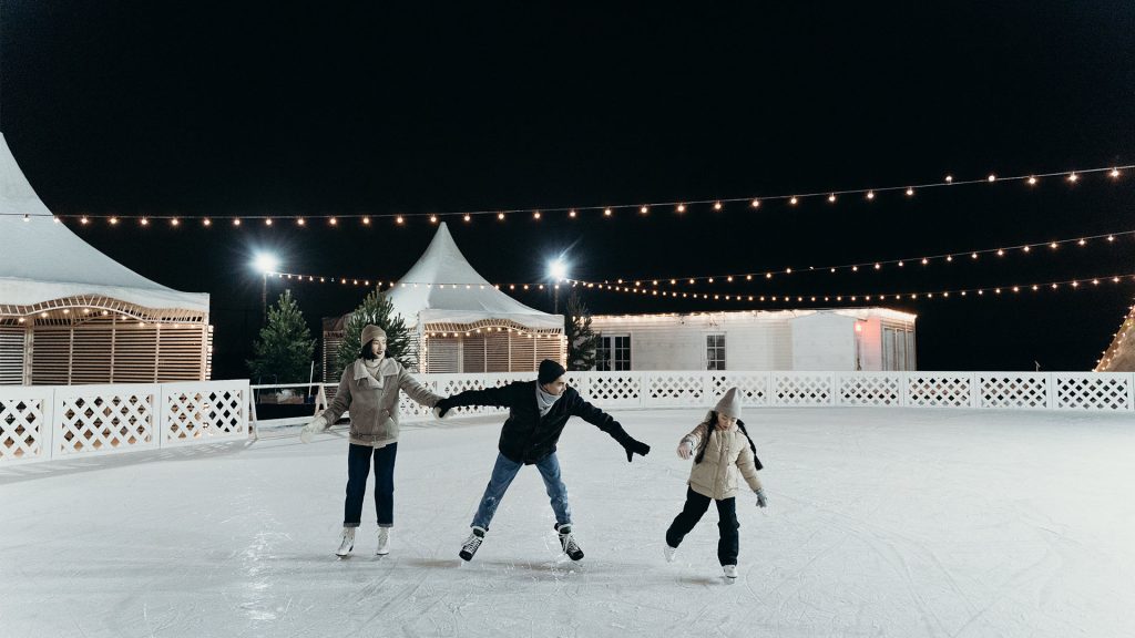 A family of three ice skating in the evening, the father in the middle reaching for their daughter