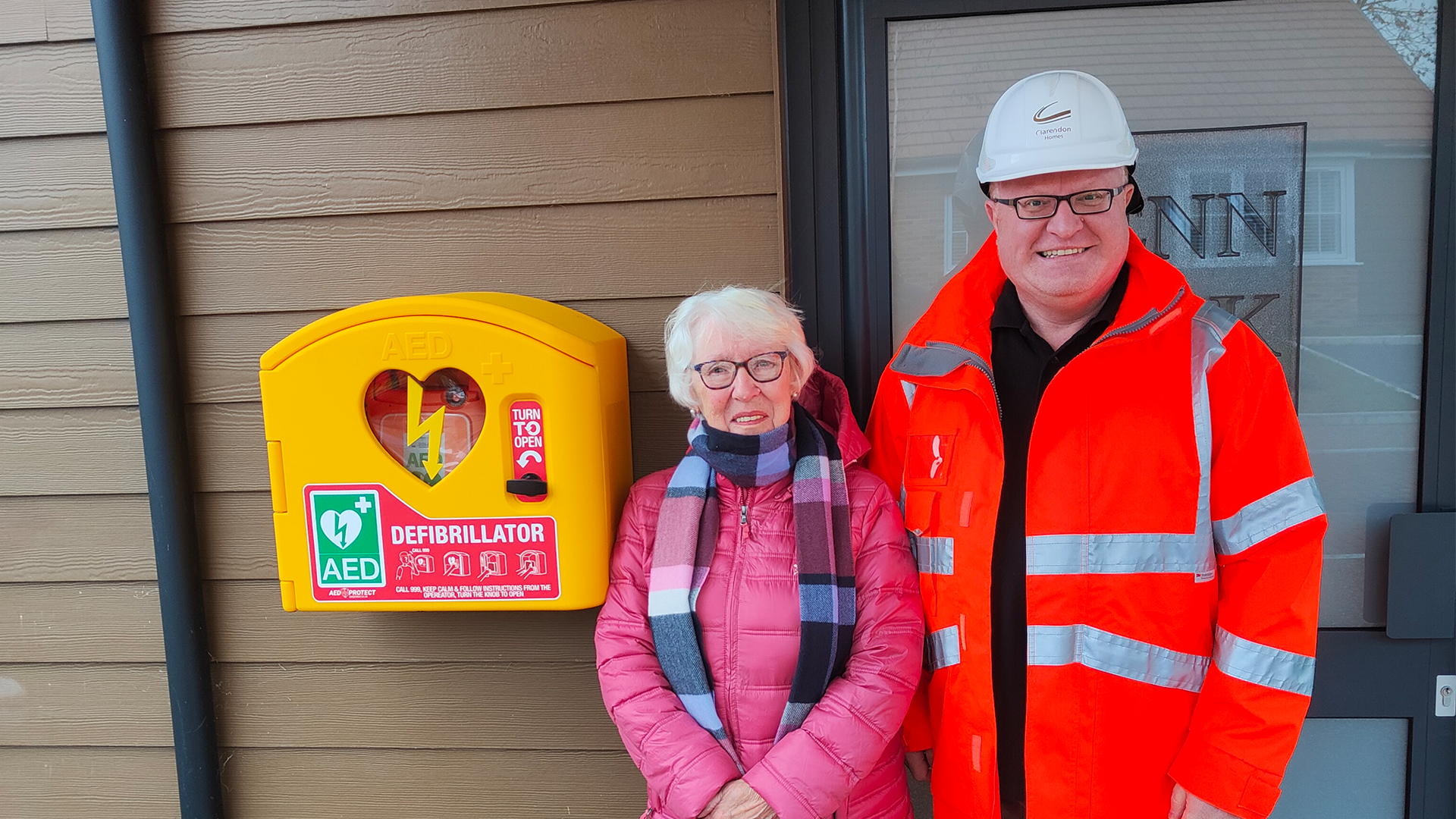 A man in a hi vis and an elderly woman stand next to a defibrillator