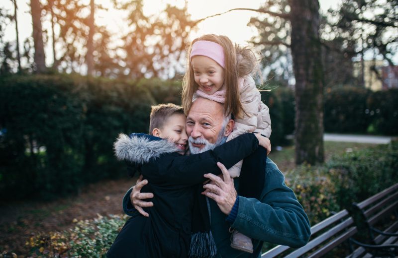 Elderly man in a park hugging his two grandchildren next to a bench.