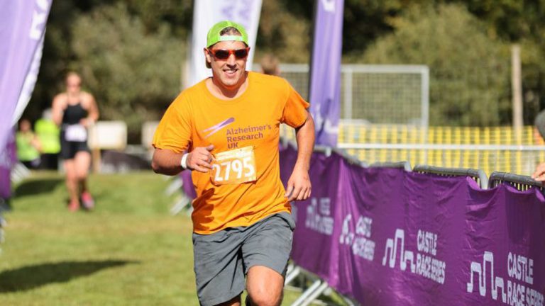 Smiling male reaching finish line of race wearing orange Alzheimer's Research UK t shirt, green hat and red sunglasses