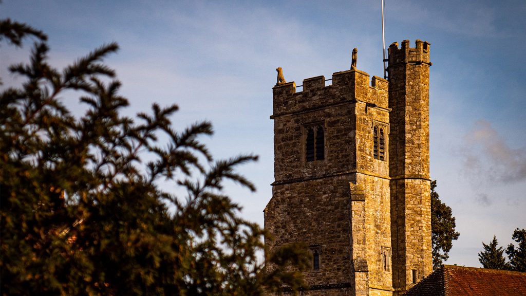 Holy Cross church tower in the sun with small gargoyles on each corner, there is an out of focus plant to the left.
