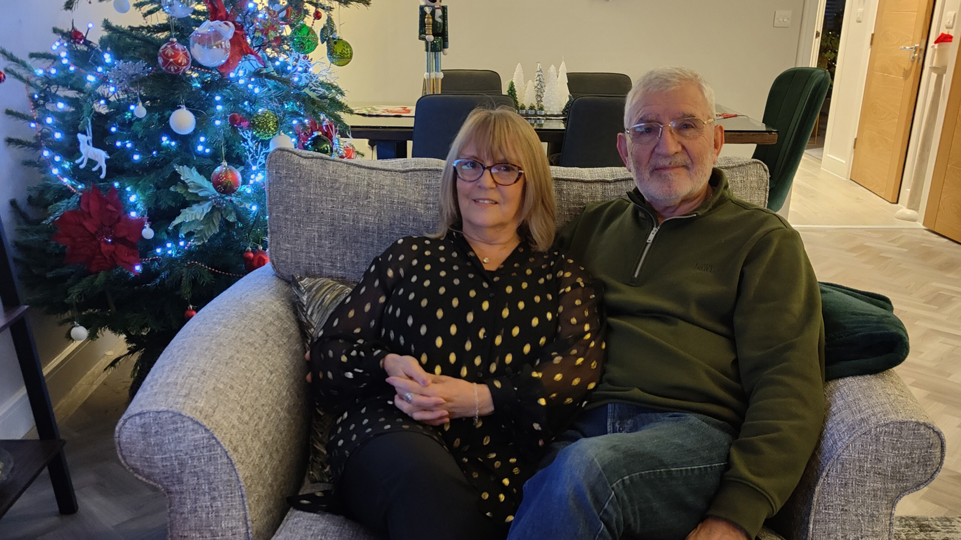 a couple sit on a loveseat while a Christmas tree illuminates the room behind them
