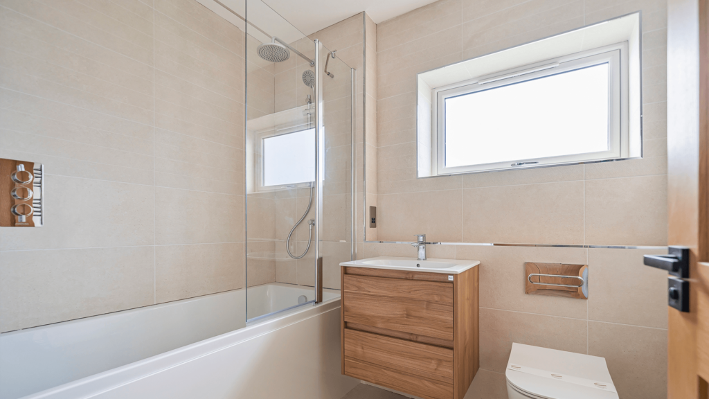 A bright bathroom with pale neutral tiling and silver accents. The bath has a glass shower door that rests open. The sink has a wooden cabinet with two drawers.