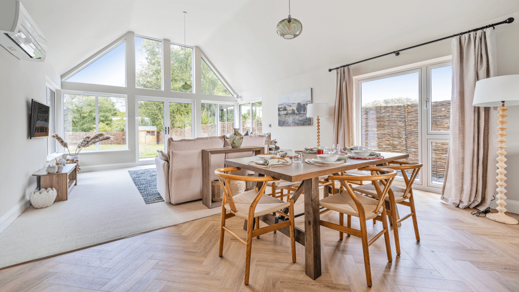 A dining/ living area with one wall full of windows and double doors at the centre. The walls and furniture is pale neutral and the dining table is a cool wood, the chairs warm with cotton woven seats.