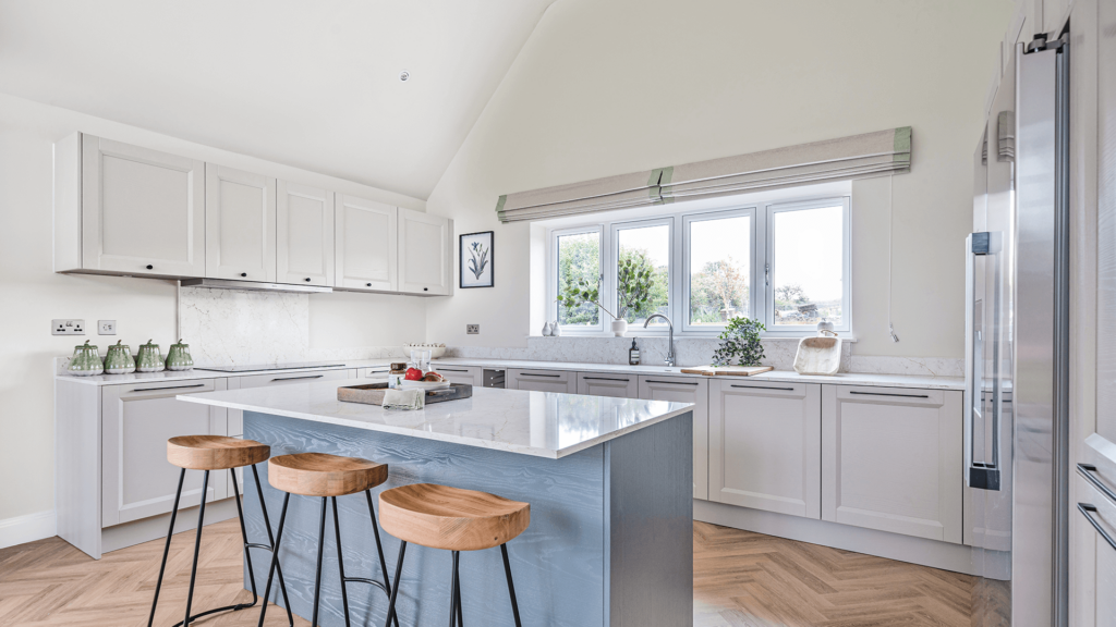 A bright airy open kitchen. The kitchen island has blue cabinets and a pale surface. Three bar stools with wooden seats sit at the island, the main kitchen cabinets have white doors and black handles. There is a long window above the sink