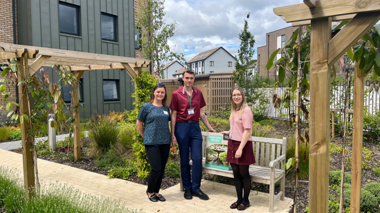 Three people stand in front of a bench in a garden- in between then is a bird bath in a box, the people on the left work at the care home.