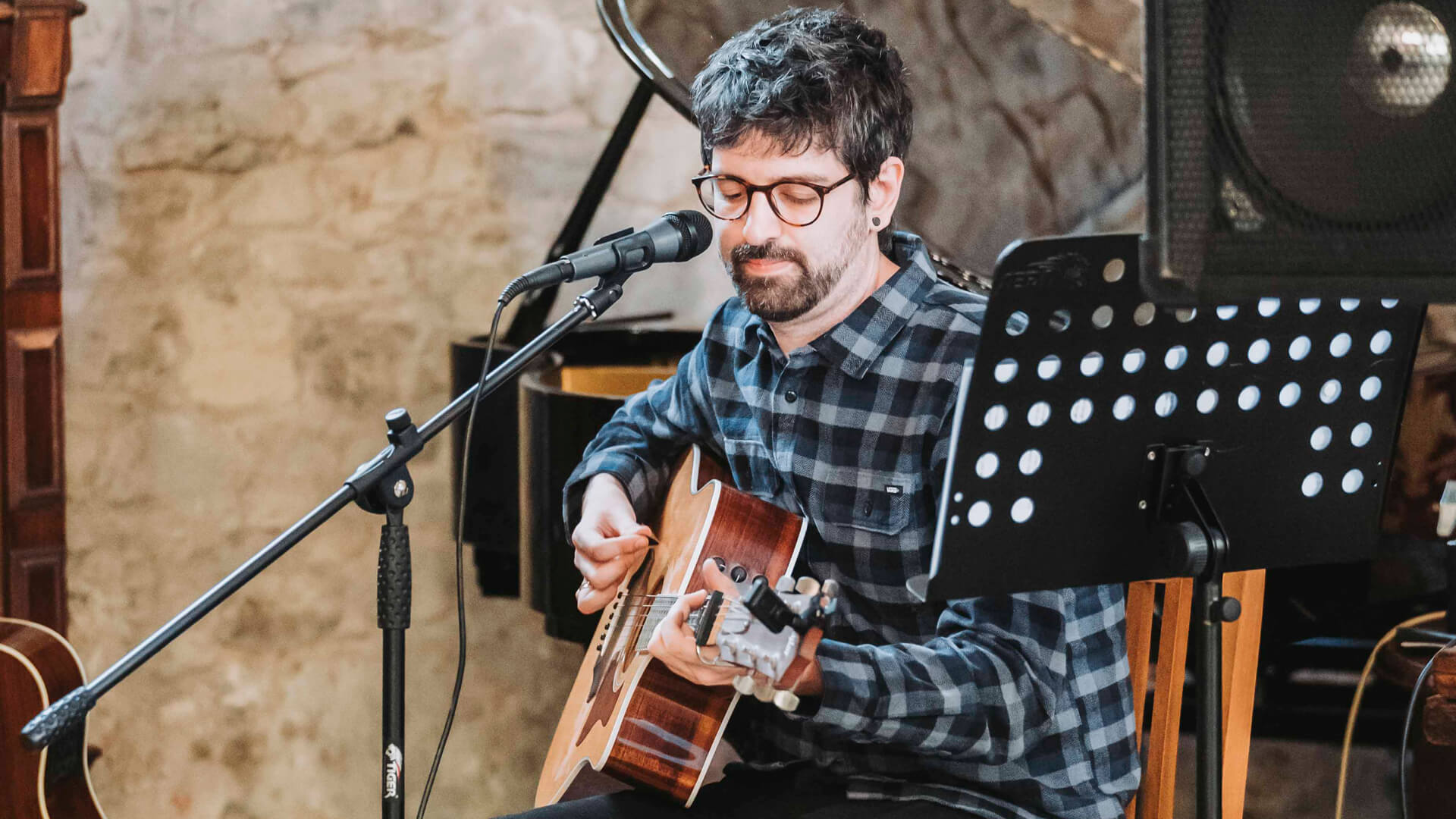 man with a bear wearing a blue chequered shit and playing a guitar with a microphone