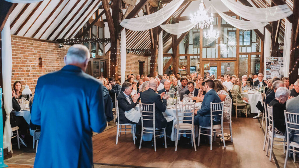 man in a dark blue suit giving a speech in front of a room full of people sitting down at tables with chandeliers hanging from the ceiling