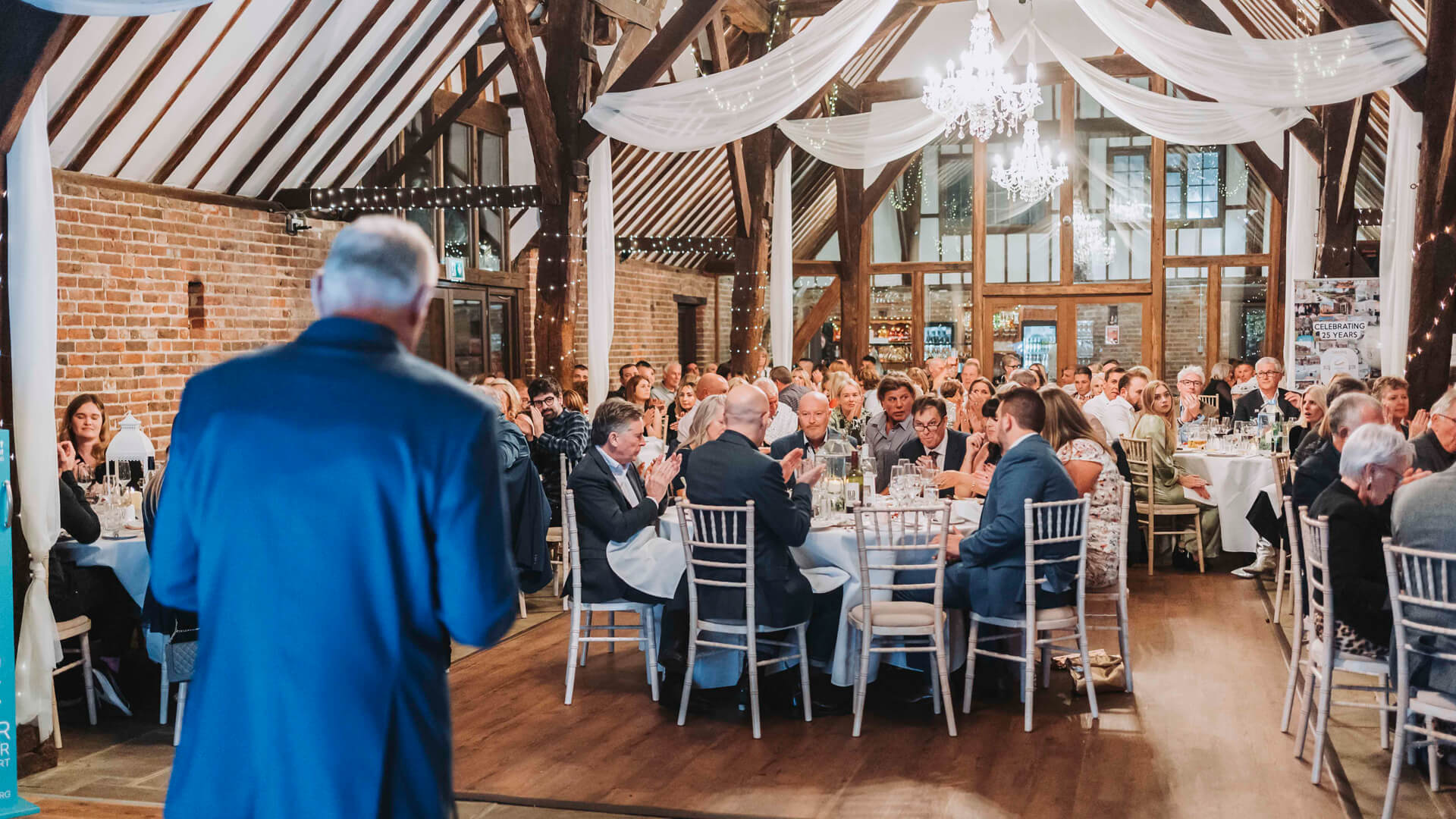 man in a dark blue suit giving a speech in front of a room full of people sitting down at tables with chandeliers hanging from the ceiling