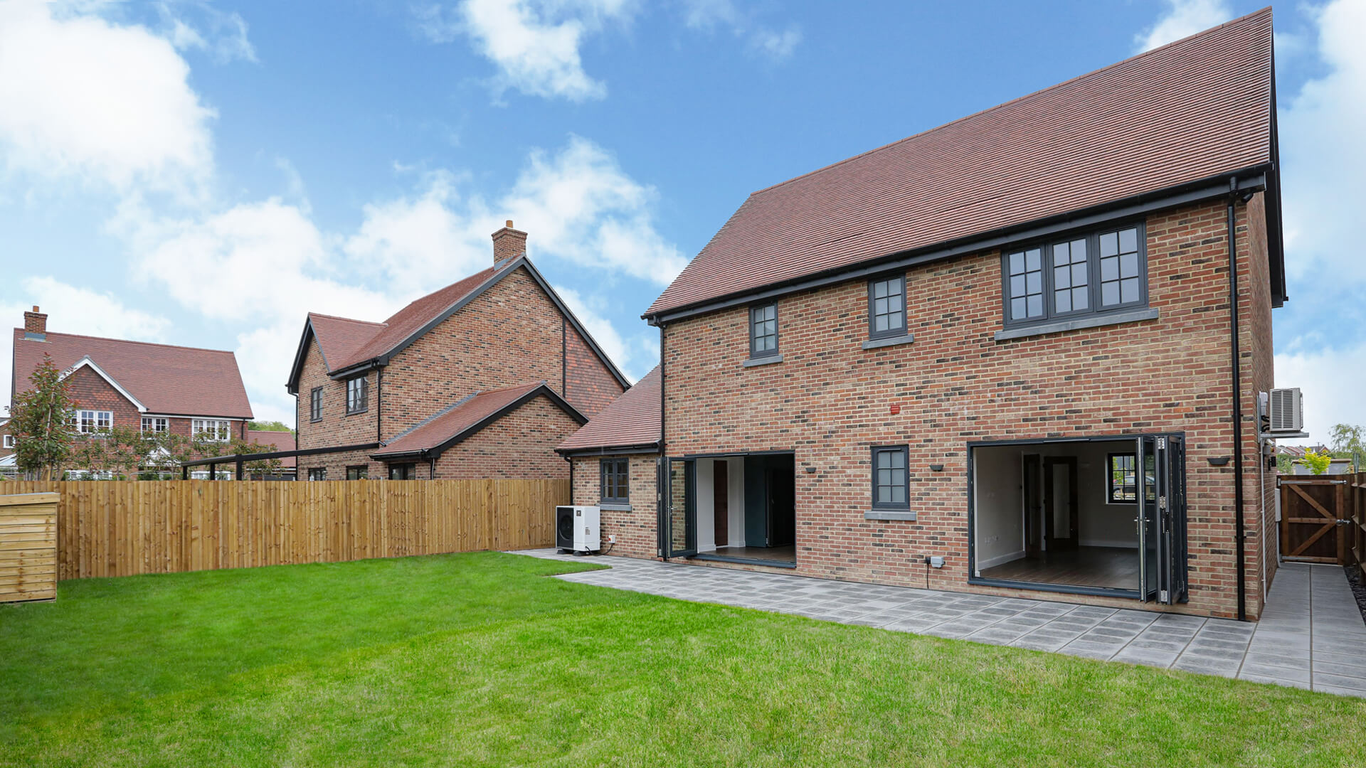 The rear of a detached house with dark bricks and grey windows and doors. Green grass and blue sky with clouds.