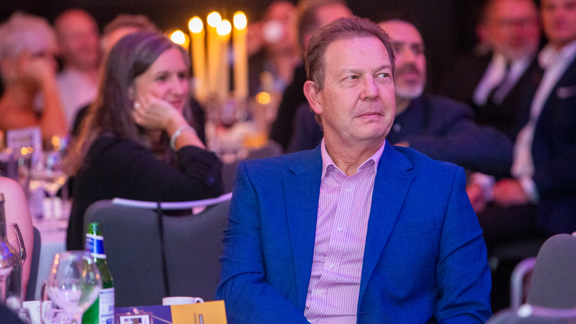 Man wearing a blue suit sitting at a table at an awards event, looking away from the camera