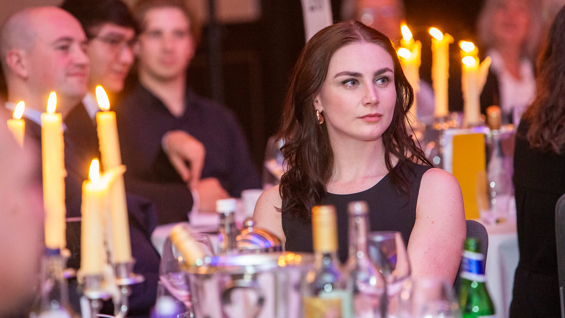 Woman with brunette hair wearing a black short sleeve dress sitting at a table with candles, at an awards event, looking away from the camera