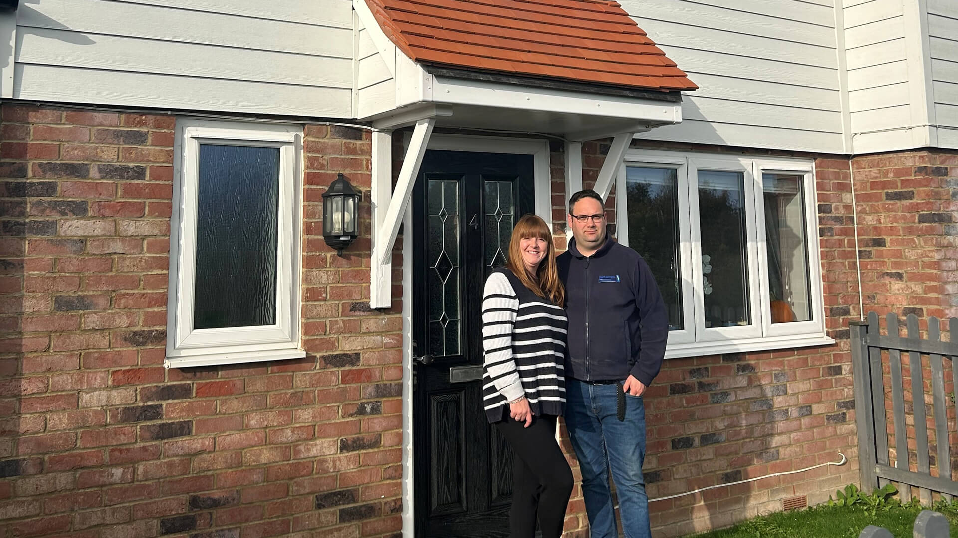 a woman and man standing posing for a photo outside a house with white cladding and a porch