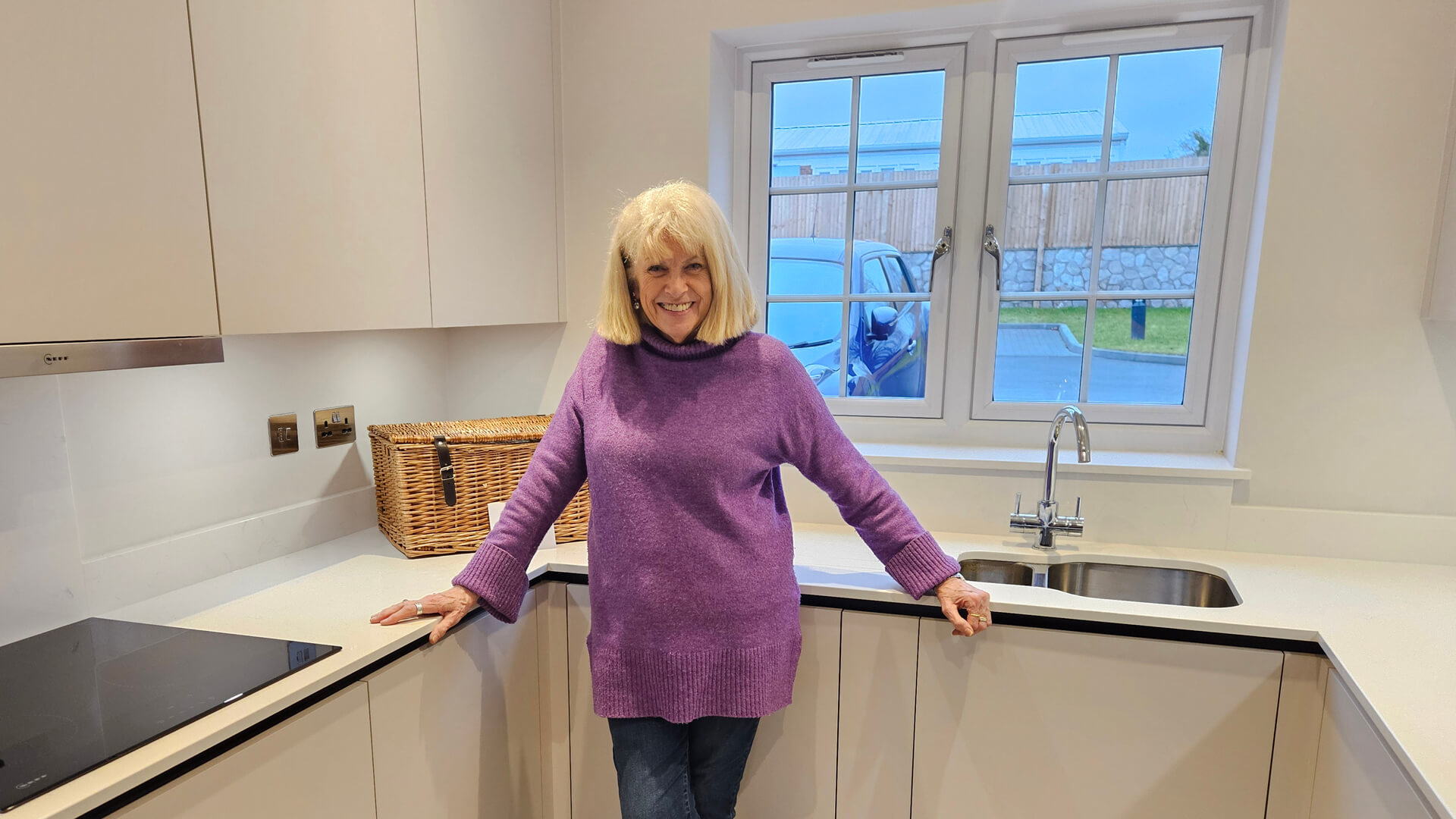 lady wearing a purple jumper and jeans posing for a photo inside a modern kitchen by the window
