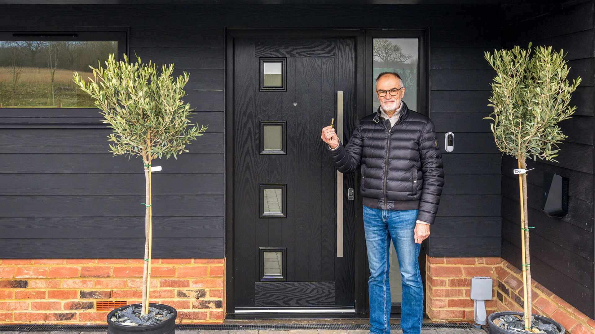 man wearing glasses and black jacket with blue jeans holding a house key outside his back front door
