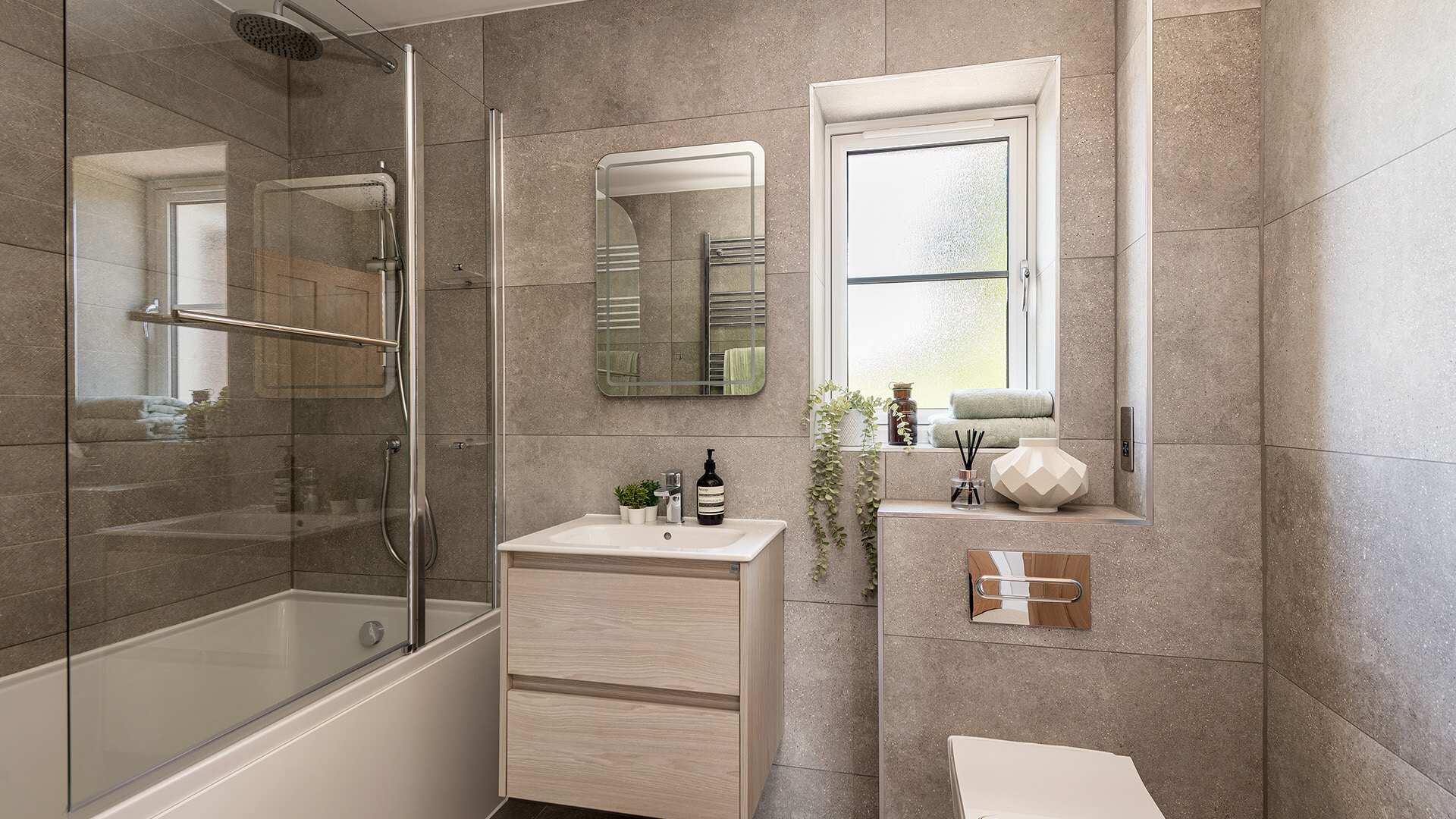 A Family bathroom with big textured matte tiles. There is a bath and shower on the left, a sink with two drawers underneath in the middle and toilet is on the right. There are plants and folded towels on the windowsill