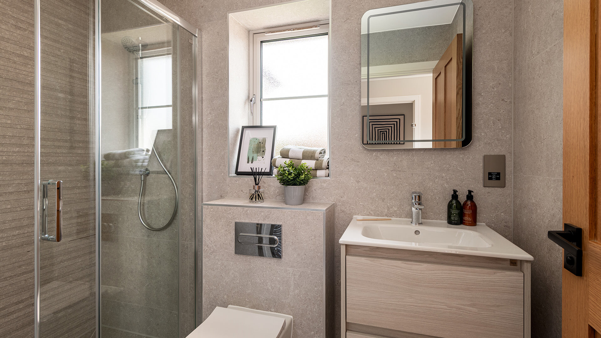 A large ensuite bathroom with big textured matte grey tiles. There is a shower on the left, a toilet in the middle and a sink with a wall mounted mirror on the right. Accessories litter the flat surfaces. Some towels, soap, a diffuser and a toothbrush