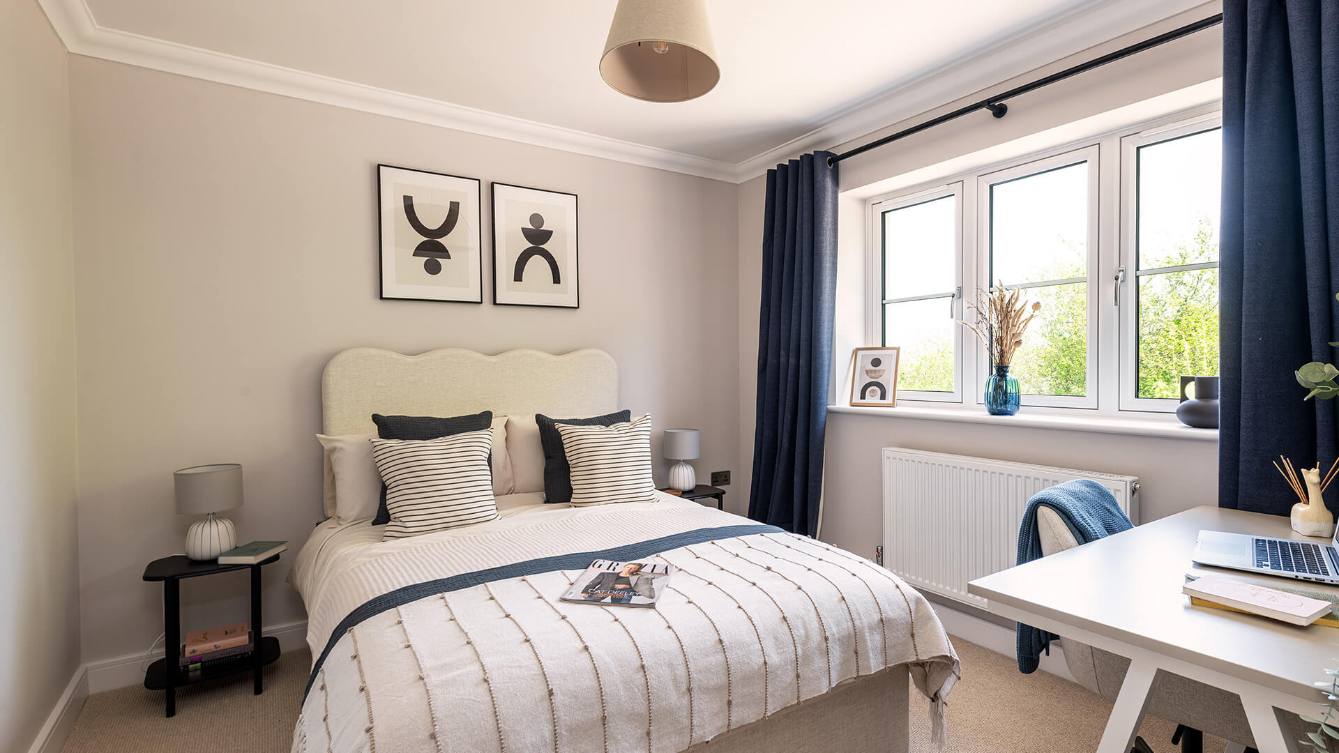 A bedroom of a show home. A double bed with white and dark blue sheets sits against the far wall, with black bedside tables and pale blue and white accent lamps. A desk sits to the right with a blue blanket over the wheelie chair.